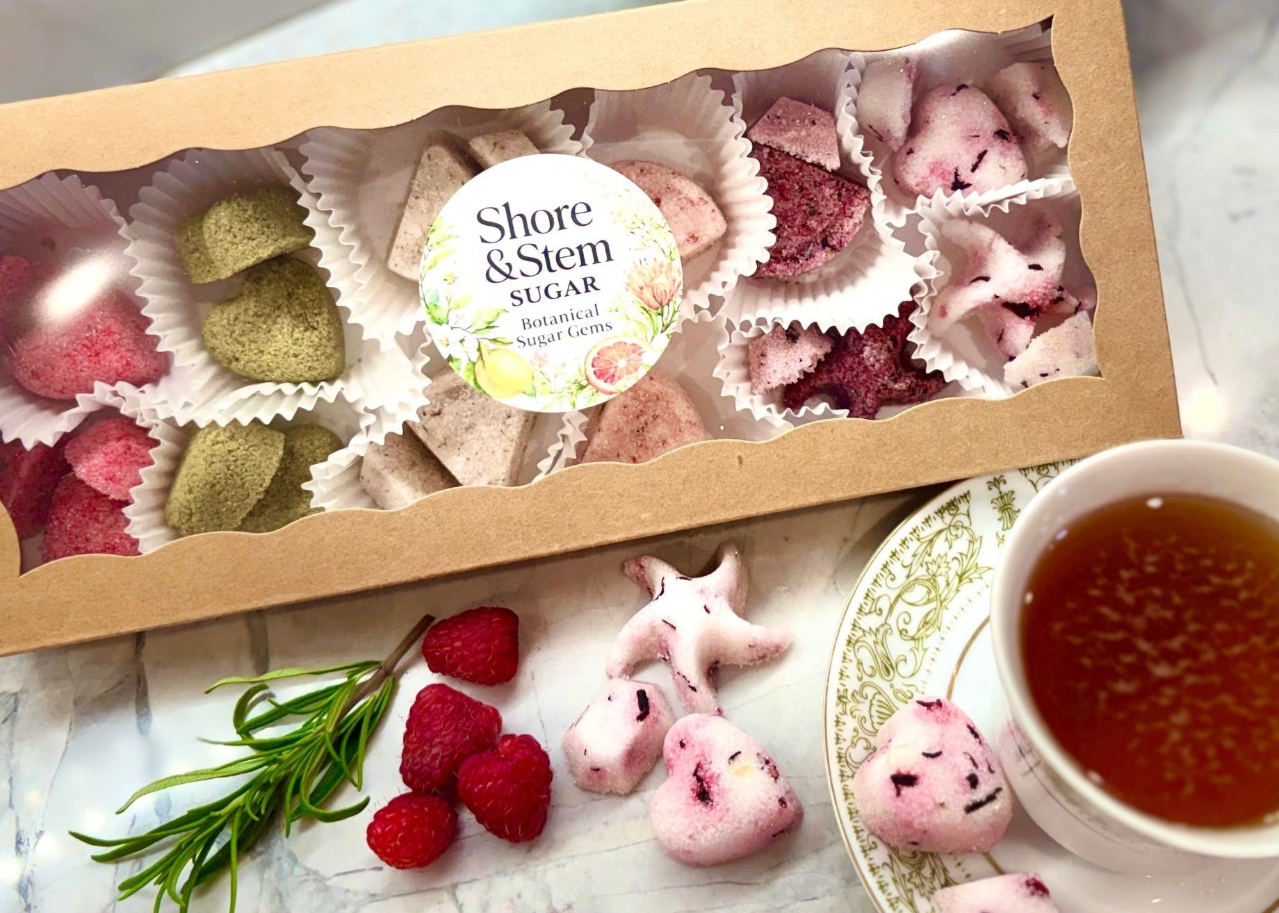Box of Shore & Stem Beverage Gems, a teacup of tea, and raspberries and Rosemary on a marble surface.